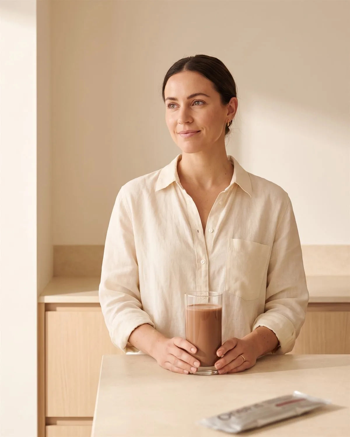A woman in a cream linen shirt at a bare stone bench in a calm, uncluttered kitchen in morning light, holding a glass of chocolate OneFit shake, with a OneFit sachet on the bench beside her